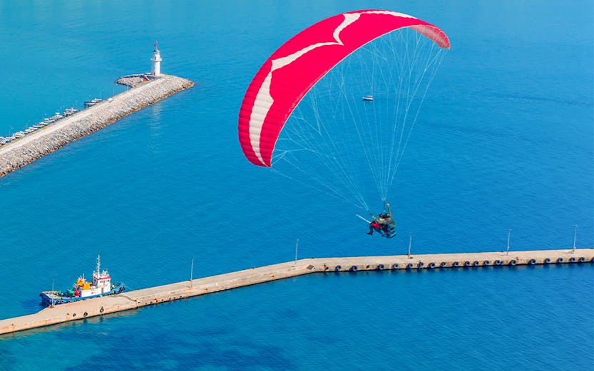 Paragliding over Alanya harbor with cityscape and lighthouse in view.