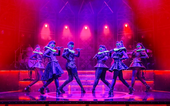 Performers on stage in vibrant costumes during the musical "Six" with dramatic lighting.