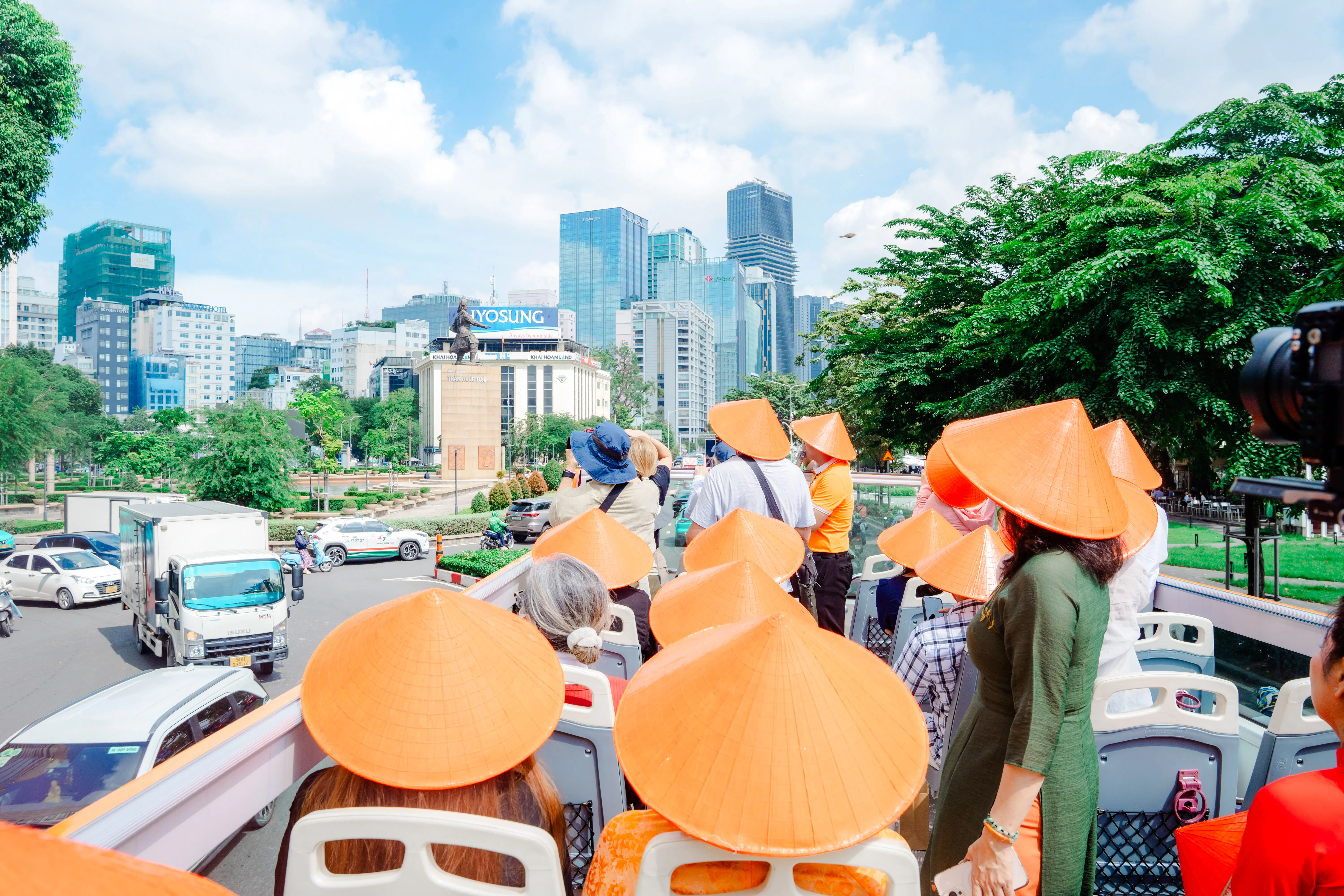 Tourists wearing conical hats on an open-top bus in Ho Chi Minh City, viewing cityscape and statue.
