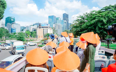 Tourists wearing conical hats on an open-top bus in Ho Chi Minh City, viewing cityscape and statue.