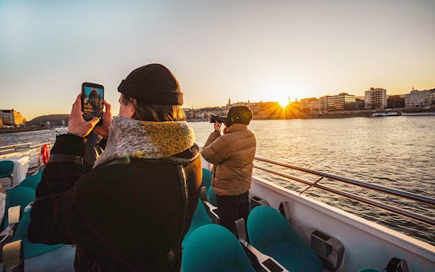 Guests photographing sunset on Budapest cruise with city skyline in view.