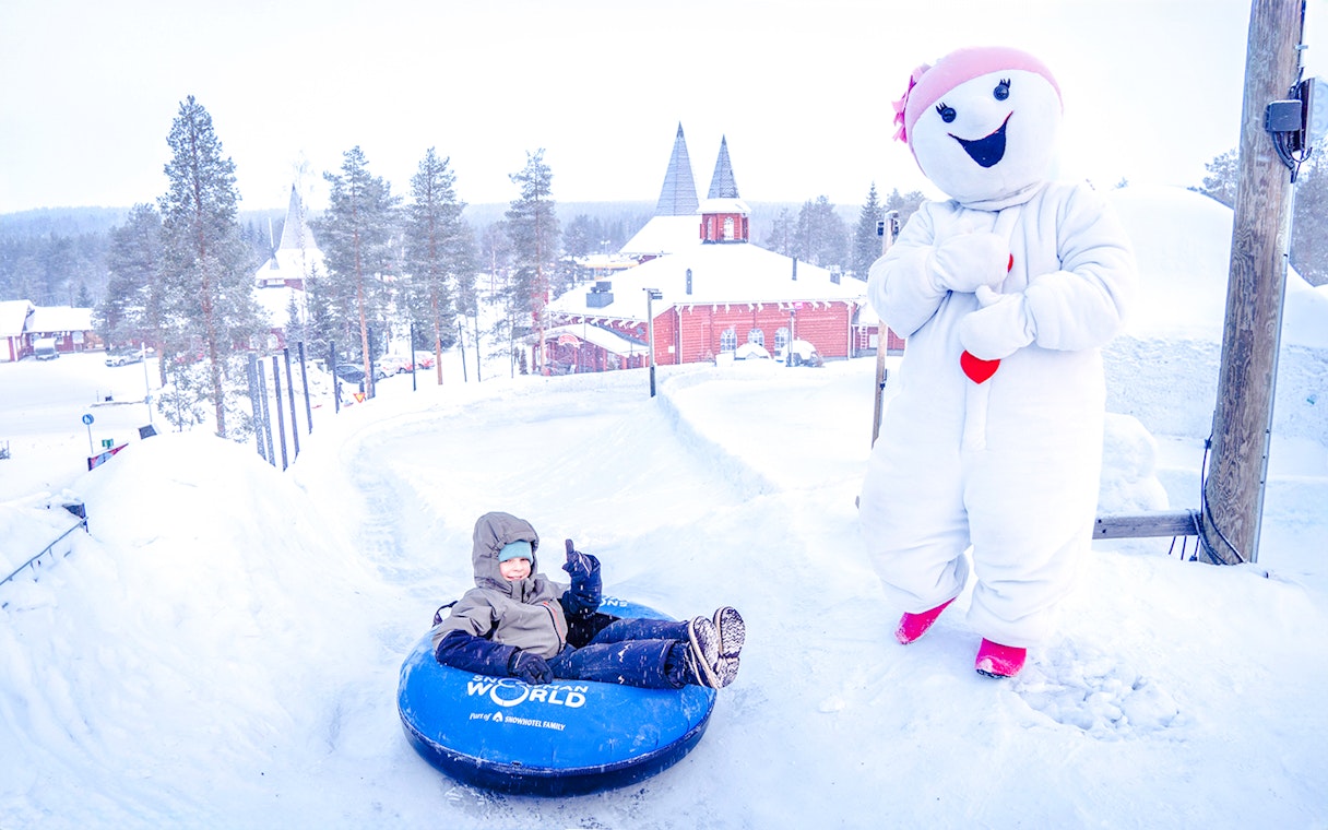 Child tubing at Snowman World, Santa Claus Village, with snowman character nearby.