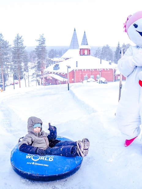 Child tubing at Snowman World, Santa Claus Village, with snowman character nearby.