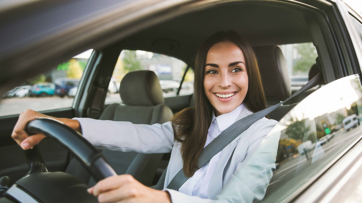 Person driving a car, smiling, with city street visible through window.