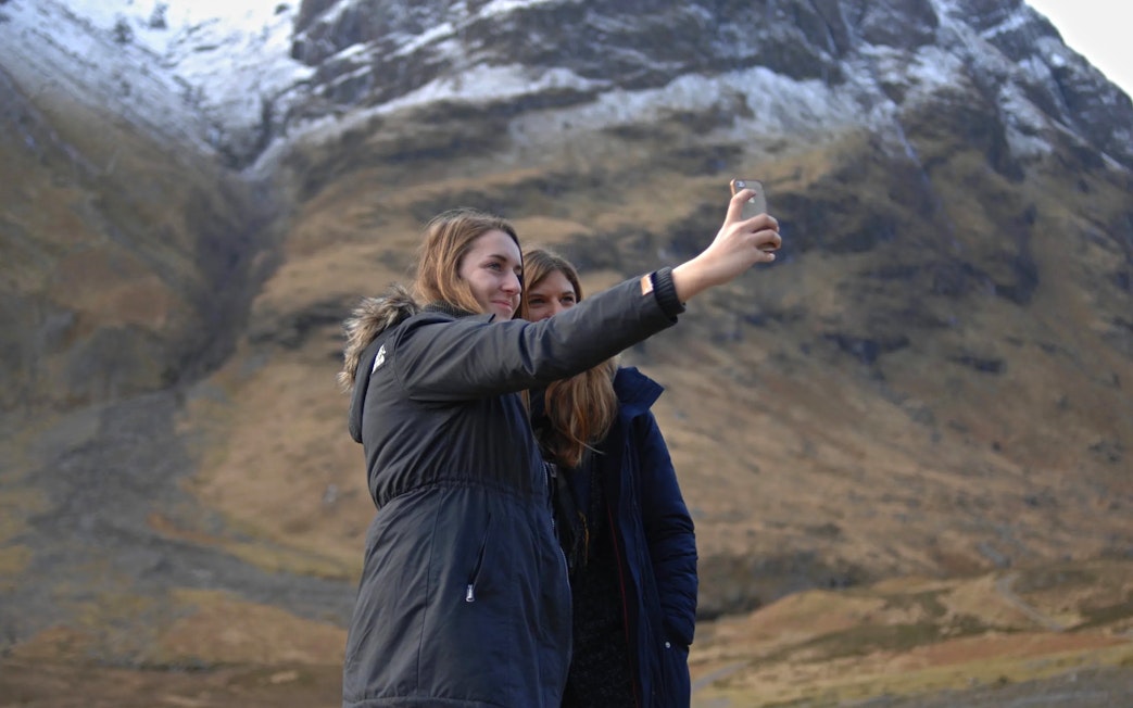 Two people taking a selfie in Glen Coe with snow-capped mountains in the background.