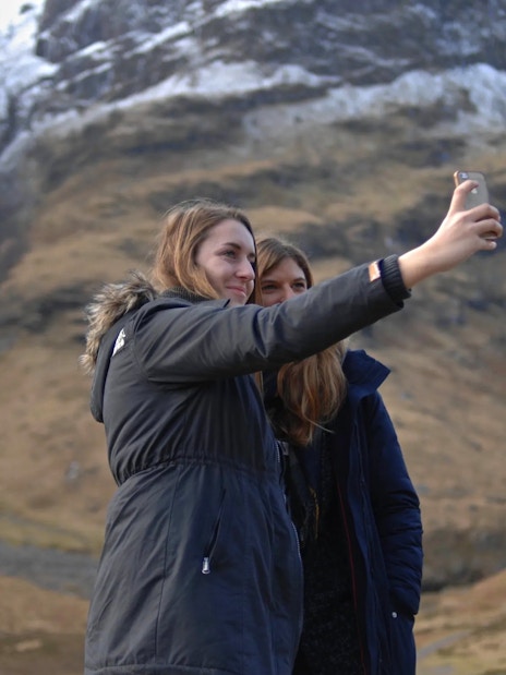 Two people taking a selfie in Glen Coe with snow-capped mountains in the background.