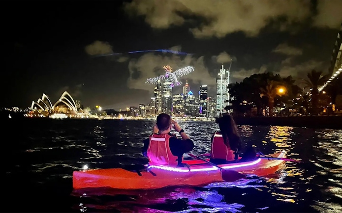 Night kayaking in LED-lit kayak towards Sydney skyline and Harbour Bridge.