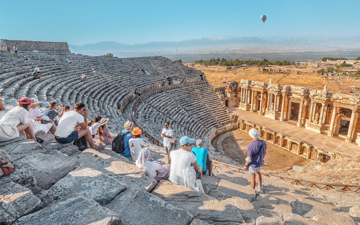 Tourists sitting in the ancient Roman theater of Hierapolis, Pamukkale, with a hot air balloon in the sky.
