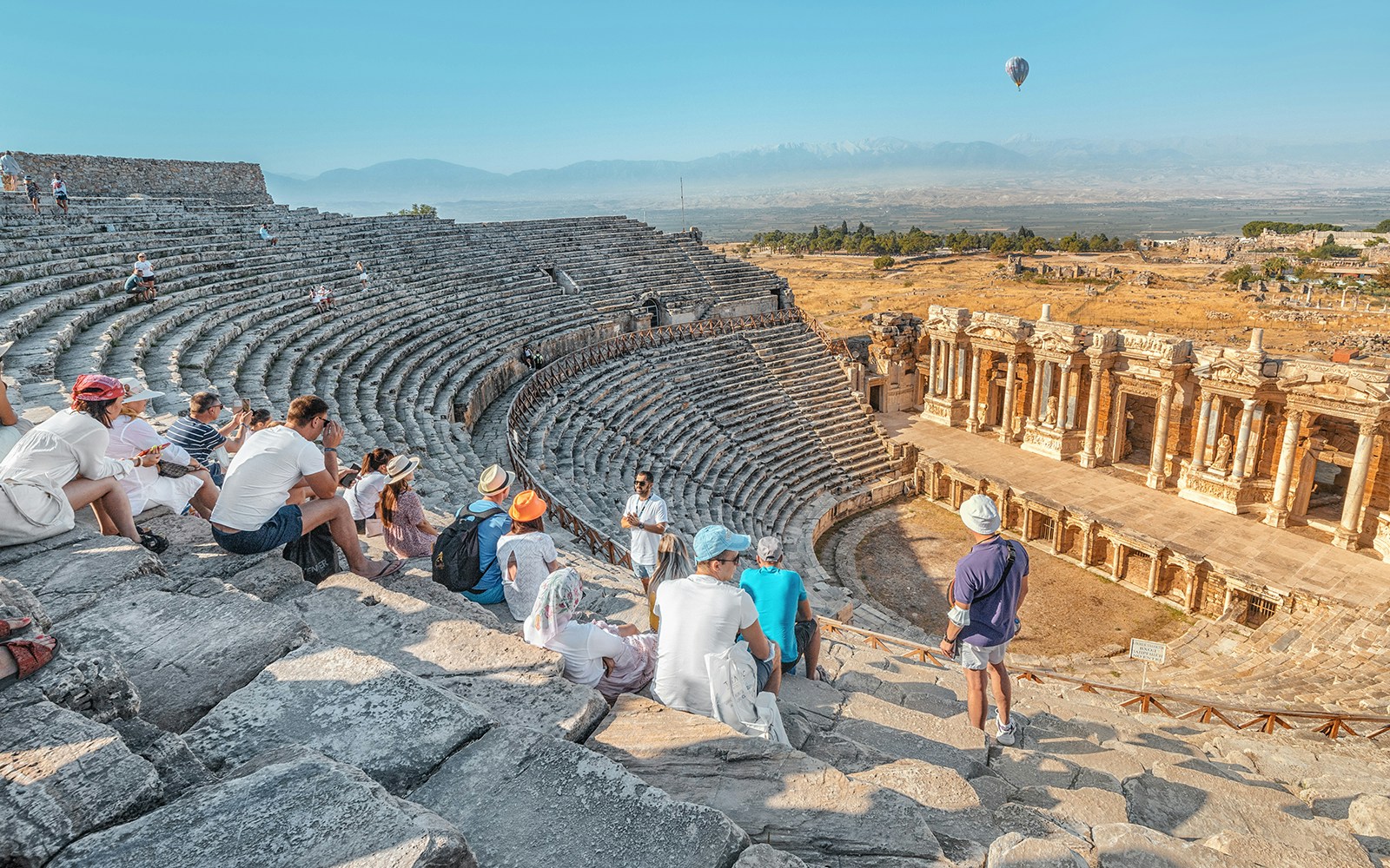 Tourists sitting in the ancient Roman theater of Hierapolis, Pamukkale, with a hot air balloon in the sky.