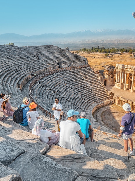 Tourists sitting in the ancient Roman theater of Hierapolis, Pamukkale, with a hot air balloon in the sky.