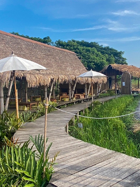 Rakna Cafe entrance with wooden walkway, thatched roof huts, and white umbrellas in Ayutthaya.