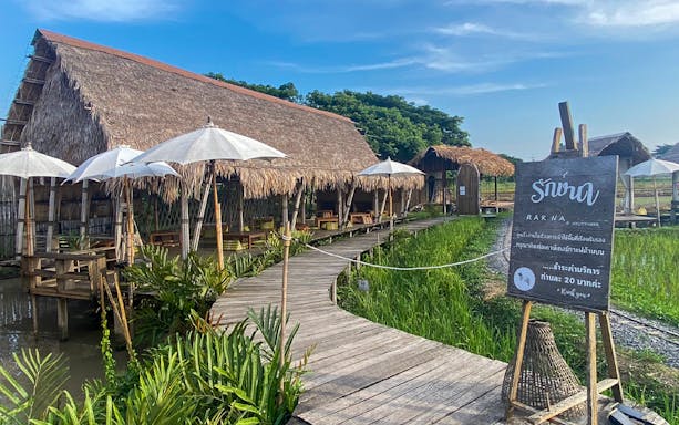 Rakna Cafe entrance with wooden walkway, thatched roof huts, and white umbrellas in Ayutthaya.