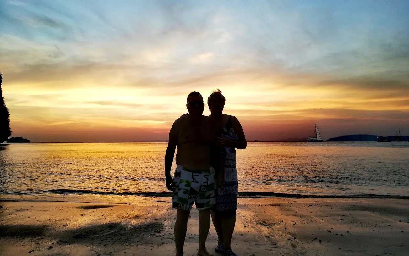 Sunset view on Krabi beach with silhouettes of two people, sailboats in the distance.