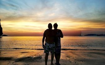 Sunset view on Krabi beach with silhouettes of two people, sailboats in the distance.