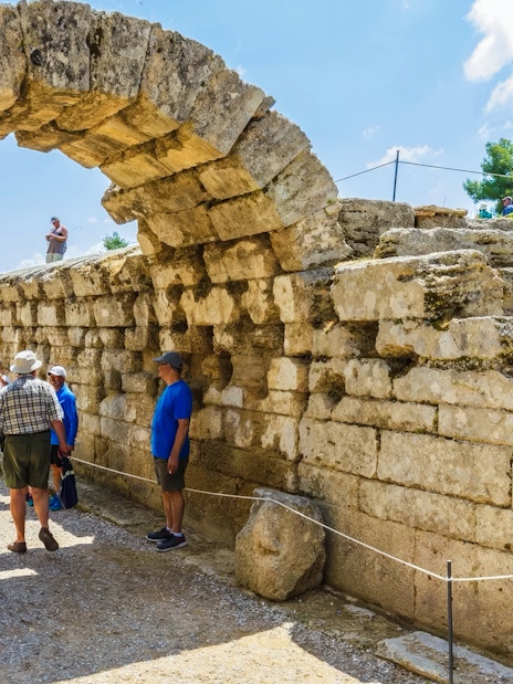 Entrance archway to ancient stadium, Olympia, Greece, with tourists exploring the site.