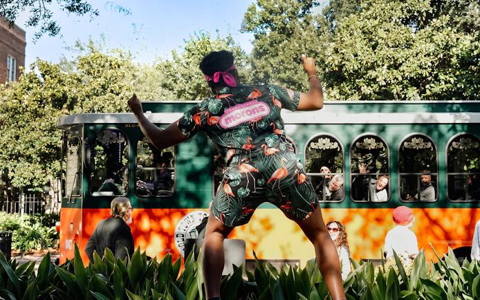 Person in colorful outfit dancing in front of Savannah trolley with passengers.
