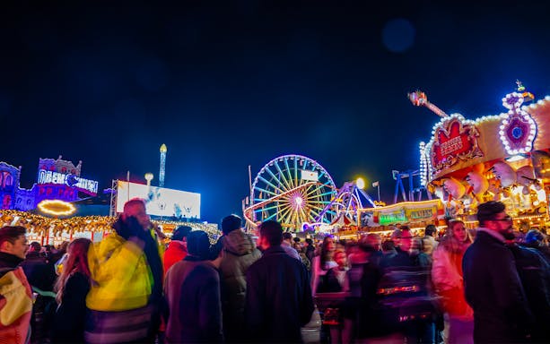 People walking at night in Hyde Park's Winter Wonderland, London, with colorful rides and attractions.