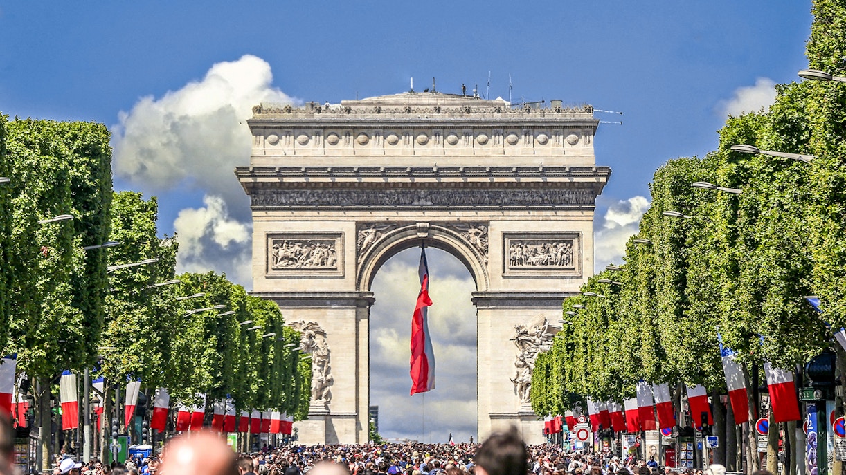 Tourists gather at the Arc de Triomphe in Paris, with French flags lining the street.