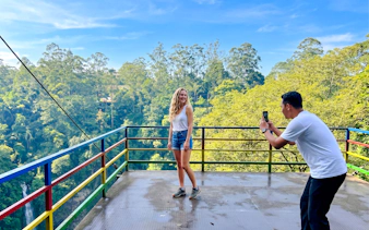 Person posing on a colorful viewing platform at Curug Cimahi, Bandung, with lush forest backdrop.