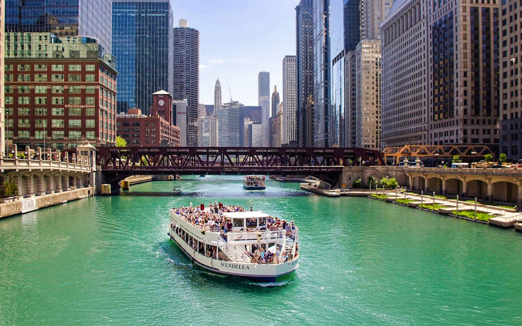 Chicago River cruise boat passing under a bridge with city skyline.