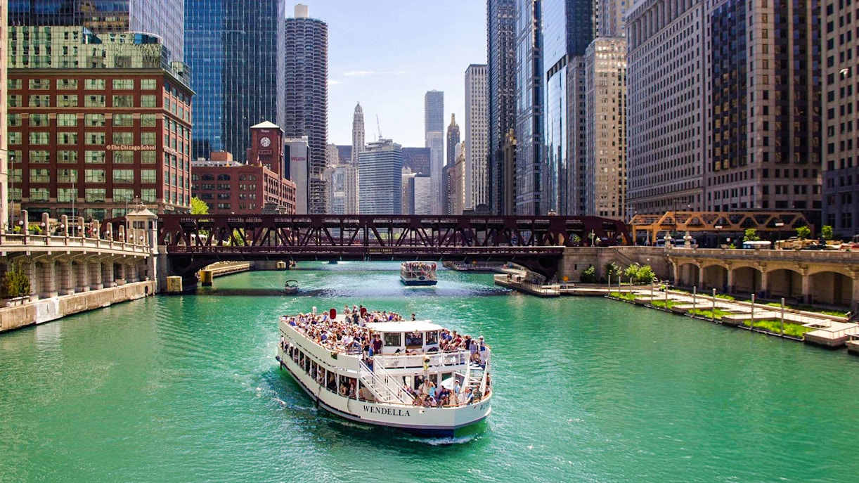 Chicago River cruise boat passing under a bridge with city skyline.