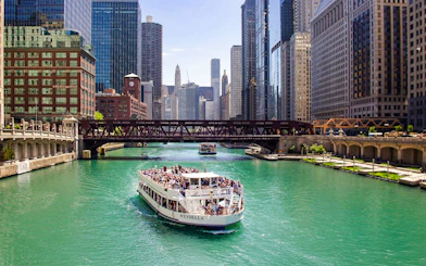 Chicago River cruise boat passing under a bridge with city skyline.