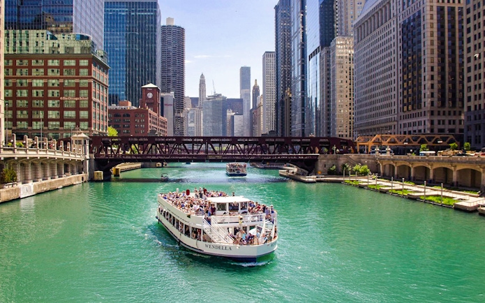 Chicago River cruise boat passing under a bridge with city skyline.