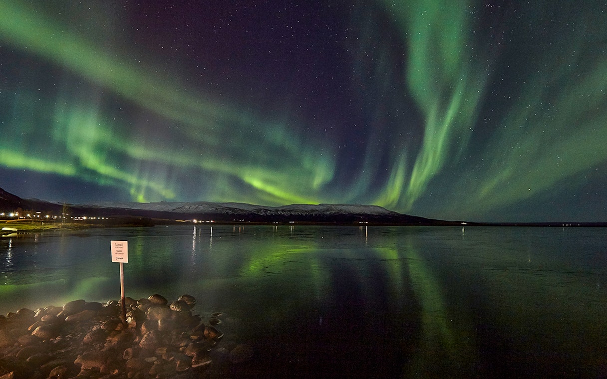 Northern Lights over a calm lake with distant snowy mountains in Iceland.
