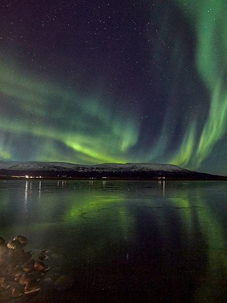 Northern Lights over a calm lake with distant snowy mountains in Iceland.