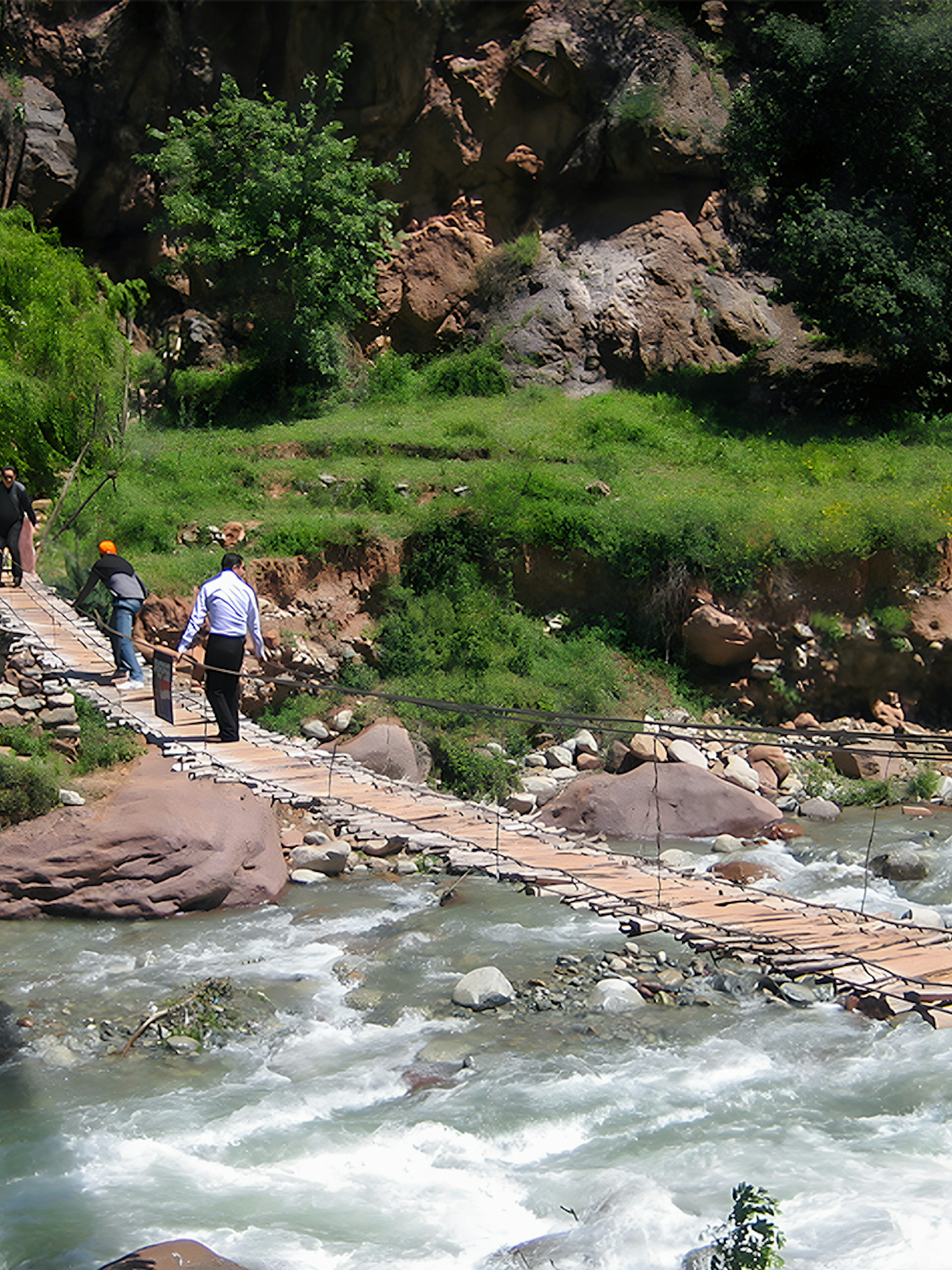 People crossing a wooden bridge over a river in a lush, rocky landscape.