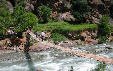 People crossing a wooden bridge over a river in a lush, rocky landscape.