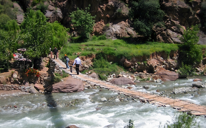 People crossing a wooden bridge over a river in a lush, rocky landscape.