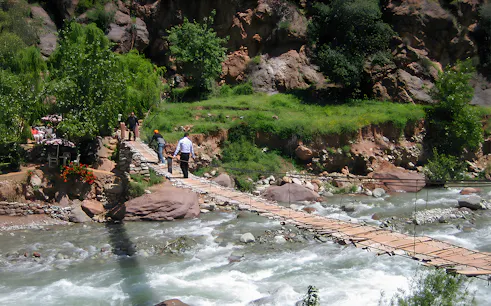 People crossing a wooden bridge over a river in a lush, rocky landscape.