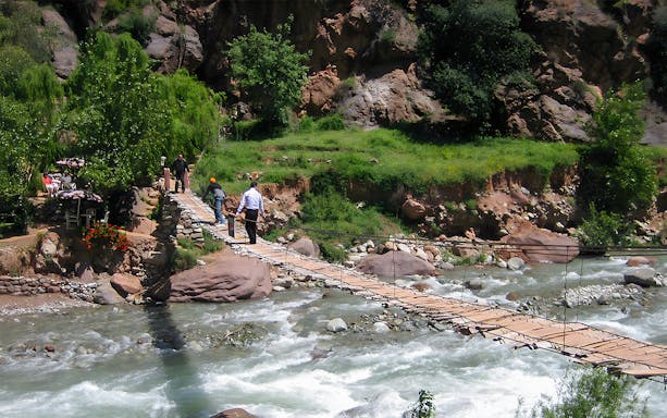 People crossing a wooden bridge over a river in a lush, rocky landscape.