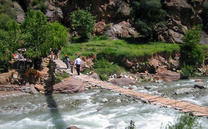 People crossing a wooden bridge over a river in a lush, rocky landscape.