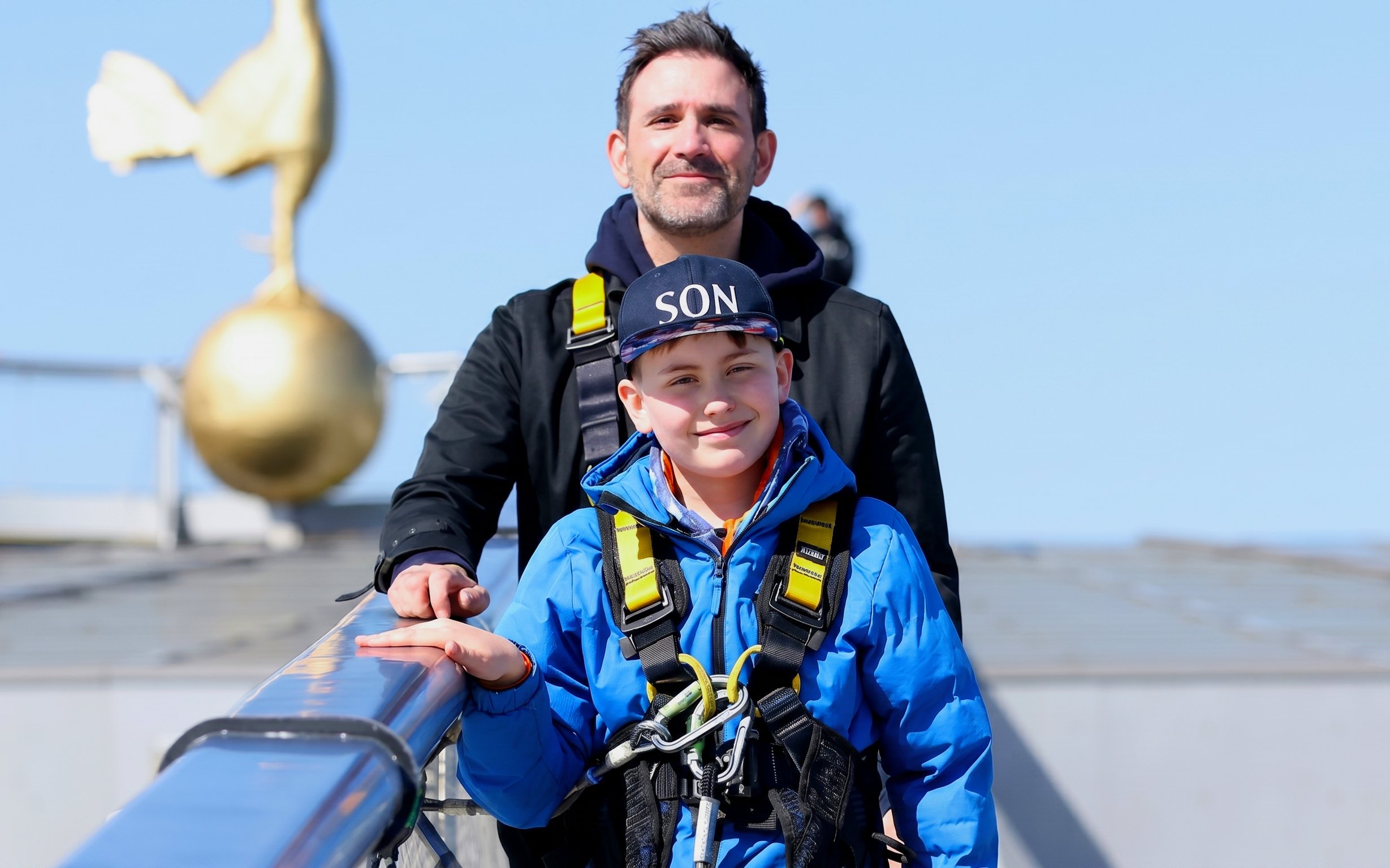 Father and son in harnesses on Dare Skywalk with golden cockerel statue in background.