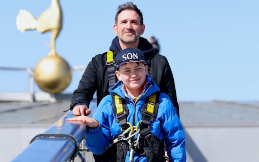Father and son in harnesses on Dare Skywalk with golden cockerel statue in background.