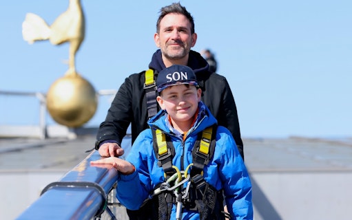 Father and son in harnesses on Dare Skywalk with golden cockerel statue in background.