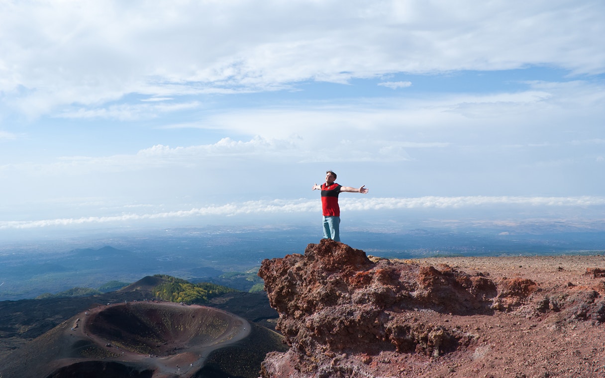 Person standing on rocky edge of Mount Etna with arms outstretched, Catania, Italy.