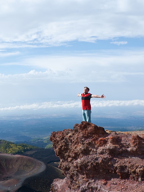 Person standing on rocky edge of Mount Etna with arms outstretched, Catania, Italy.
