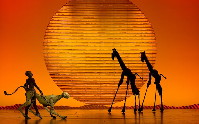 Performers in "The Lion King" musical with animal puppets against a sunset backdrop.
