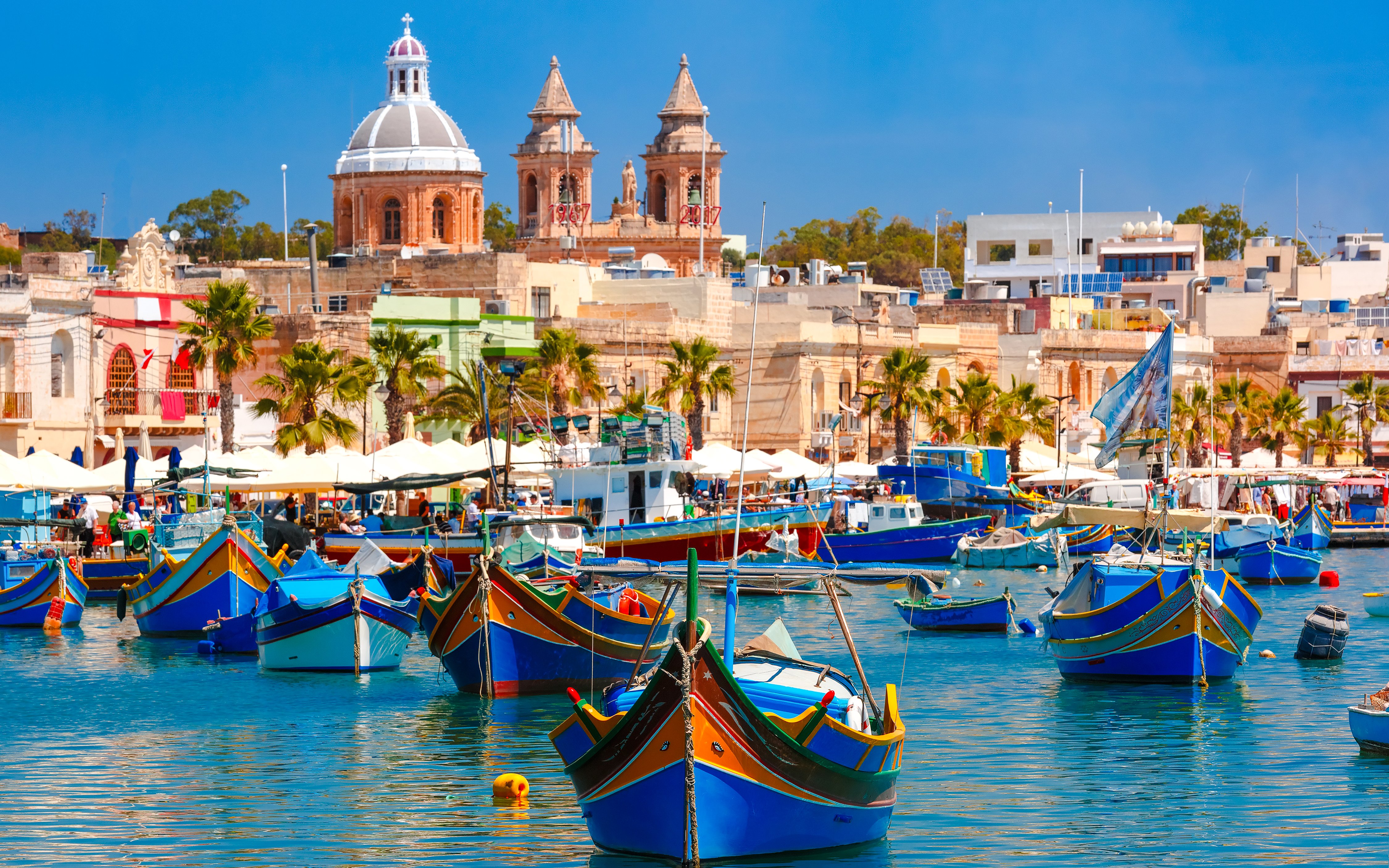 Traditional colorful boats in Marsaxlokk Harbor, Malta with village and church in background.