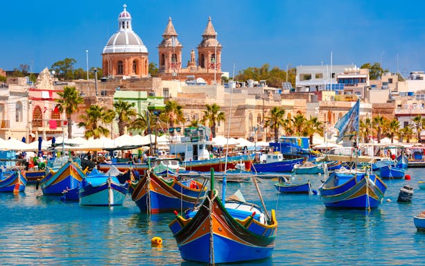 Traditional colorful boats in Marsaxlokk Harbor, Malta with village and church in background.