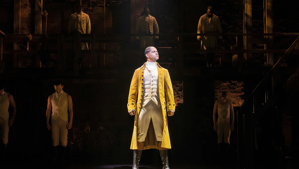 Hamilton West End performance with lead actor in yellow coat on stage.