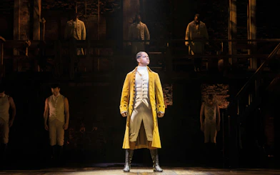 Hamilton West End performance with lead actor in yellow coat on stage.