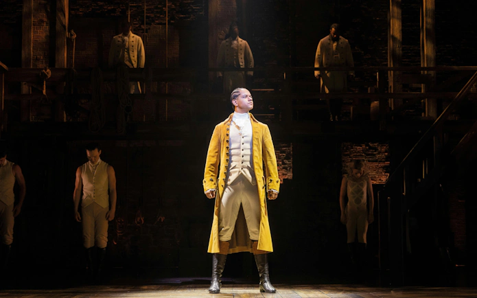 Hamilton West End performance with lead actor in yellow coat on stage.