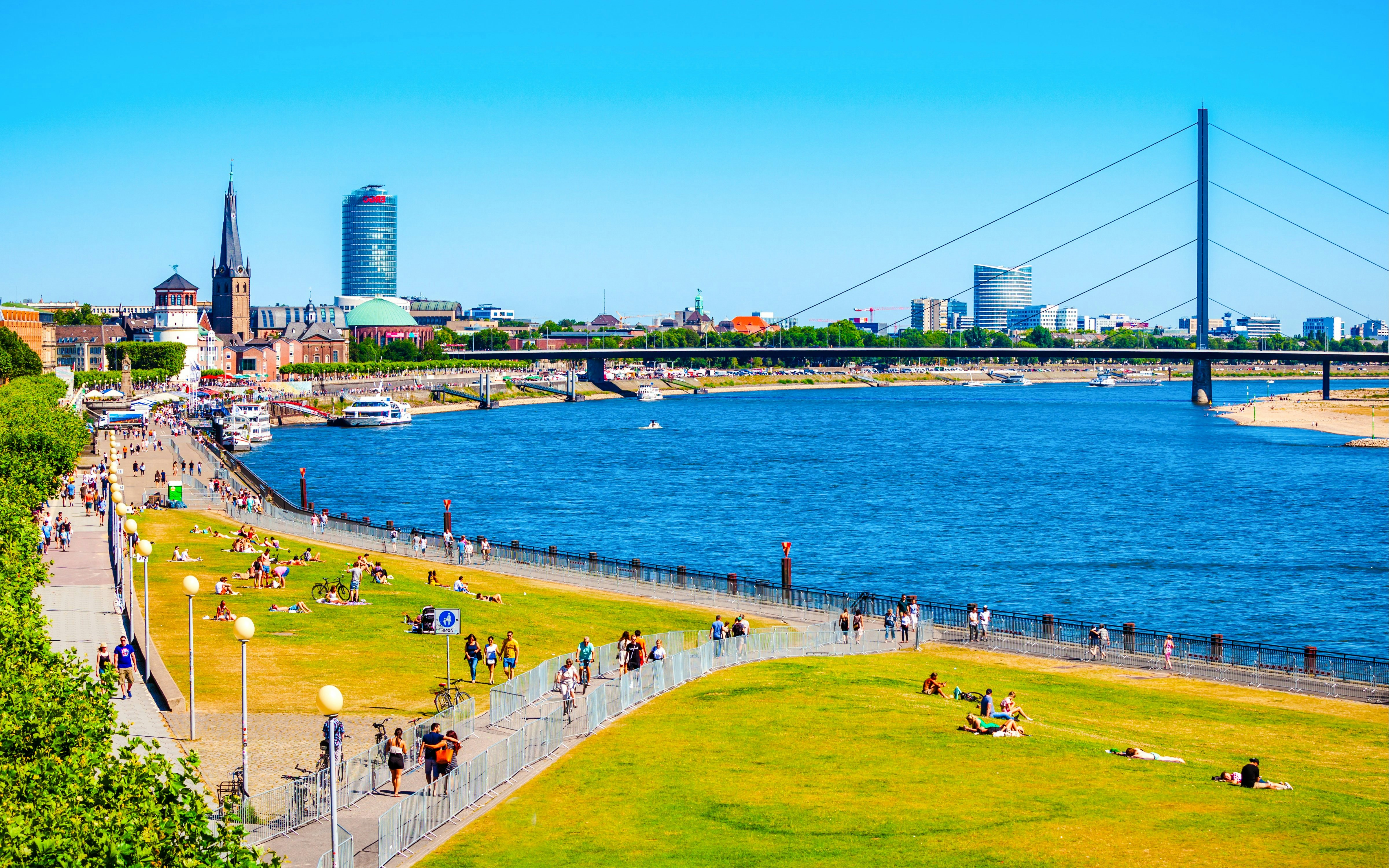 People enjoying a sunny day along the Rhine Promenade in Düsseldorf with cityscape views.