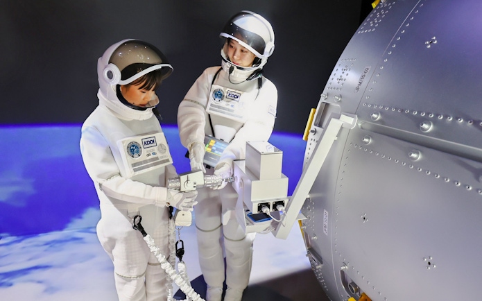 Children in astronaut suits interacting with a spaceship module at KidZania Fukuoka.