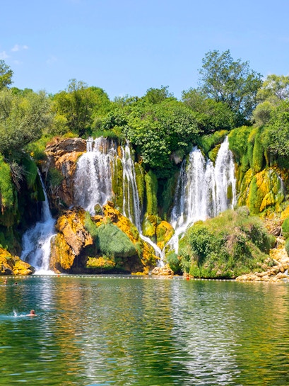 Kravica Waterfall in Bosnia with lush greenery and people swimming in the clear water.