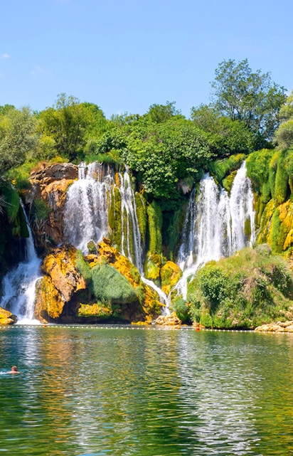 Kravica Waterfall in Bosnia with lush greenery and people swimming in the clear water.